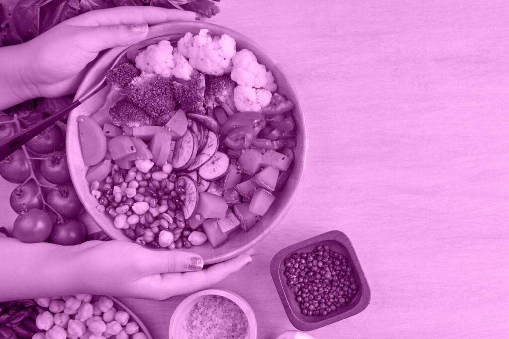 Duas mãos segurando uma cesta com frutas sobre uma mesa. Ao lado da cesta há alguns potes com outros alimentos.