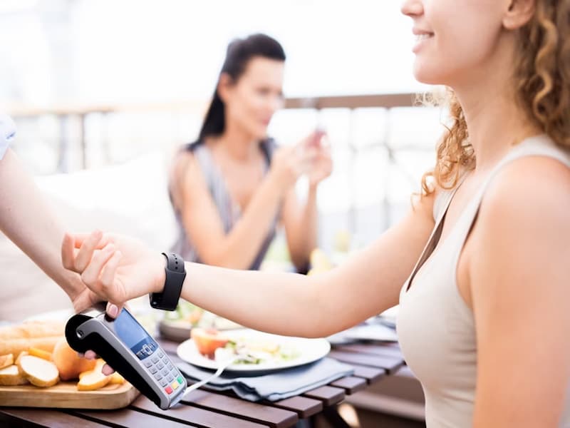 A imagem mostra uma cena que pode estar relacionada ao Programa de Alimentação do Trabalhador (PAT). Nela, um homem está realizando um pagamento com um cartão, aproximando-o de uma maquininha de pagamento móvel, possivelmente em um restaurante ou estabelecimento de alimentação.
