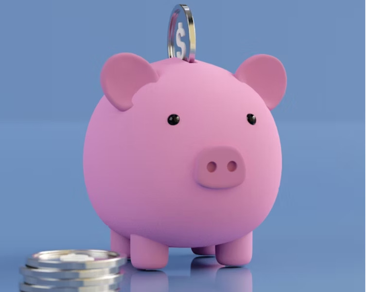 A imagem mostra um cofrinho rosa em formato de porquinho, com uma moeda com símbolo de cifrão ($) sendo inserida no topo. O fundo é azul e minimalista, e há algumas moedas adicionais colocadas ao lado do cofre, sugerindo o ato de economizar ou acumular dinheiro.