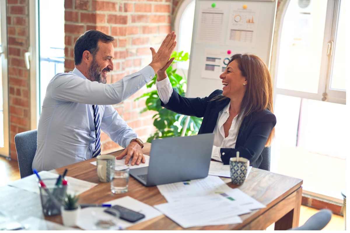 A imagem mostra dois profissionais em um ambiente de escritório celebrando um momento de sucesso com um entusiasmado "high five" (toca aqui).