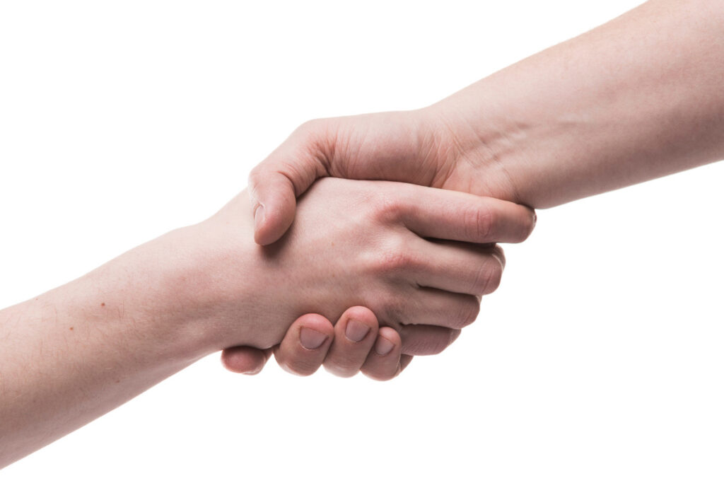 A imagem mostra duas pessoas se cumprimentando com um aperto de mão. A mão esquerda, em primeiro plano, está sendo segurada pela mão direita, que está em segundo plano.
