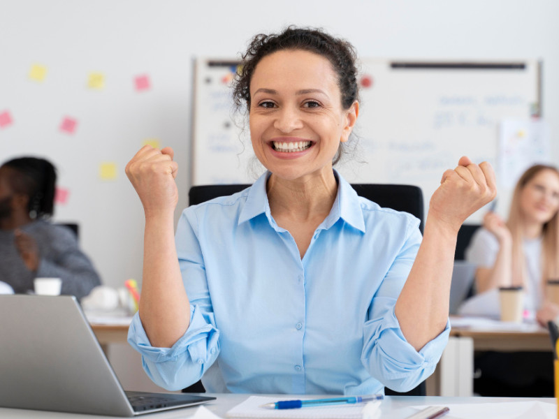 A imagem é um retrato de uma mulher em um ambiente de escritório, celebrando um sucesso ou conquista com grande entusiasmo.