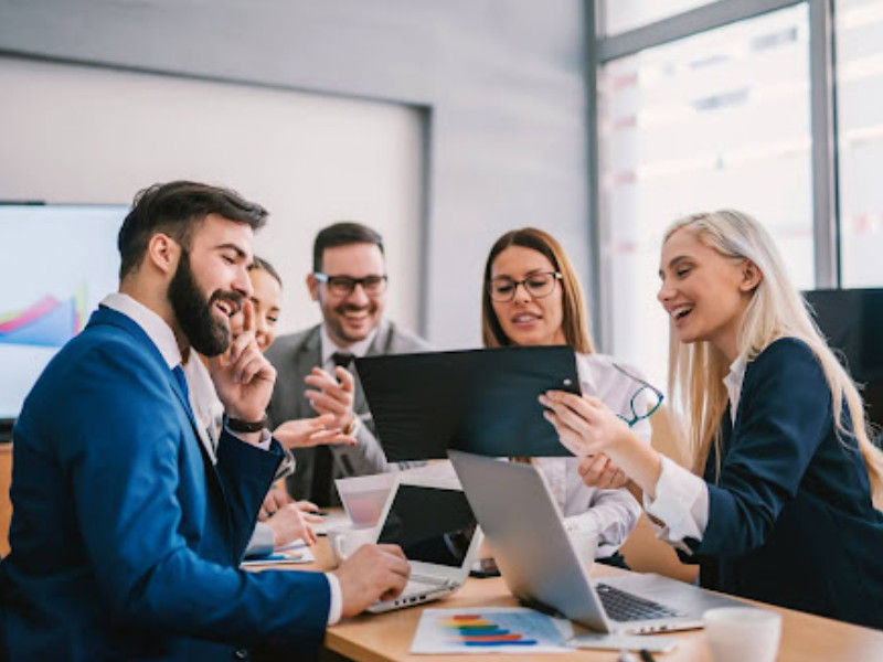 A imagem retrata um grupo de cinco profissionais de negócios em uma reunião de trabalho ou sessão de brainstorming em um escritório moderno.