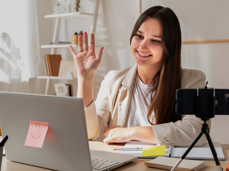A imagem retrata uma jovem profissional participando de uma videochamada, reunião online, ou gravando um vídeo a partir de um home office bem iluminado.