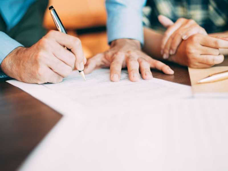 A imagem mostra um close-up das mãos de duas pessoas sobre uma mesa, analisando e preenchendo documentos. Uma das pessoas segura uma caneta e parece estar assinando um papel, enquanto a outra aponta ou segura outro documento ao lado.