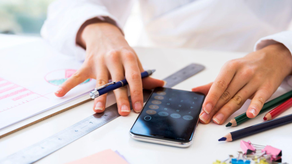 A imagem mostra um close nas mãos de uma pessoa trabalhando sobre uma mesa, realizando cálculos e organizando materiais.