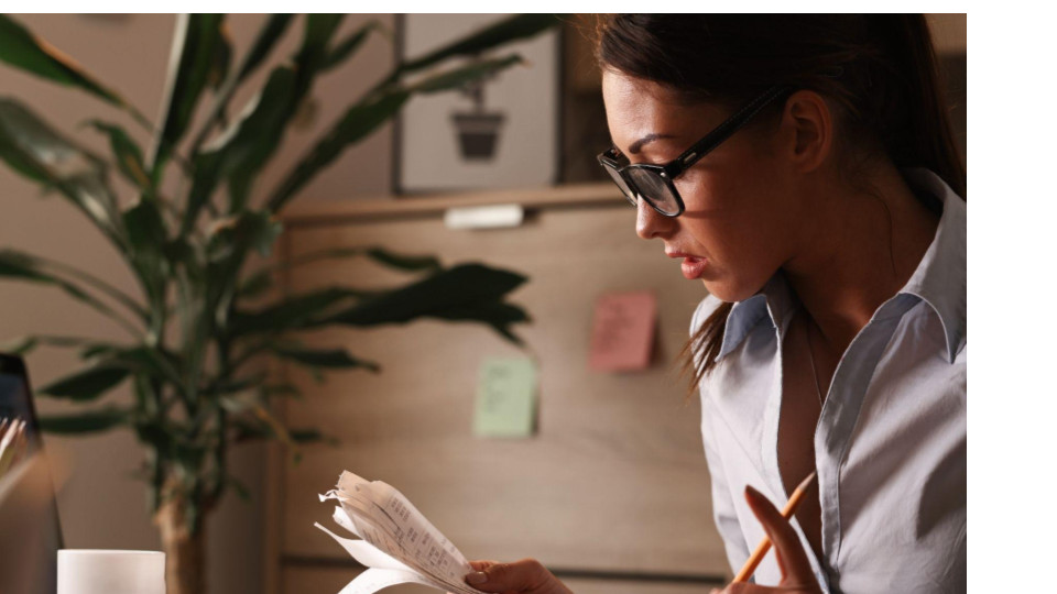 A imagem mostra uma mulher em um ambiente de escritório ou home office, analisando atentamente alguns papéis ou recibos que segura nas mãos, enquanto também segura um lápis, como se estivesse conferindo ou anotando informações.