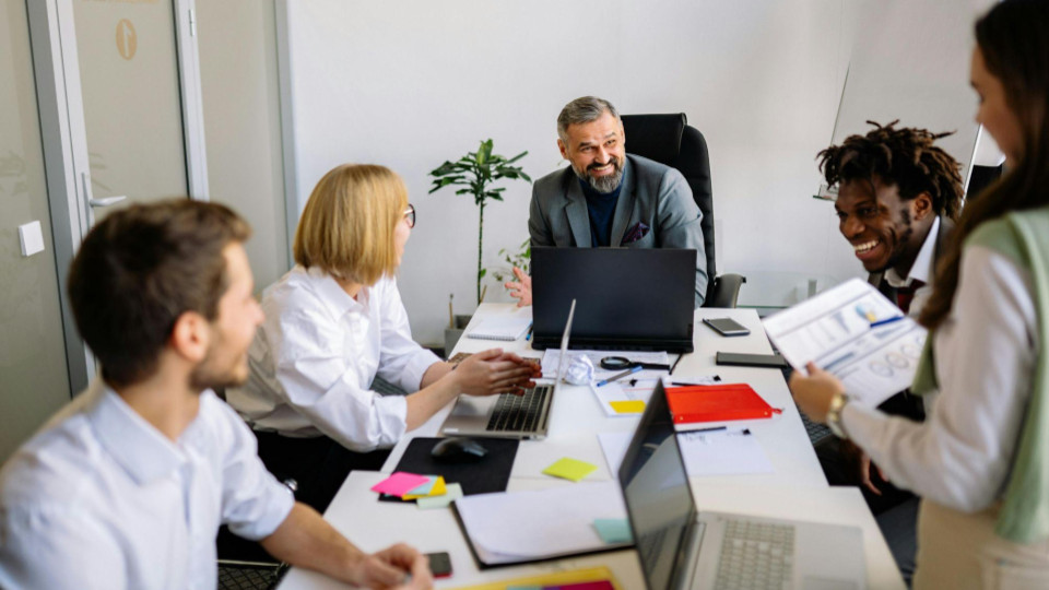 No centro da mesa, vemos um homem mais maduro, de barba grisalha e terno cinza, com uma expressão muito sorridente e acolhedora. Ele parece estar liderando a conversa de forma leve, possivelmente compartilhando um feedback positivo ou uma boa notícia.