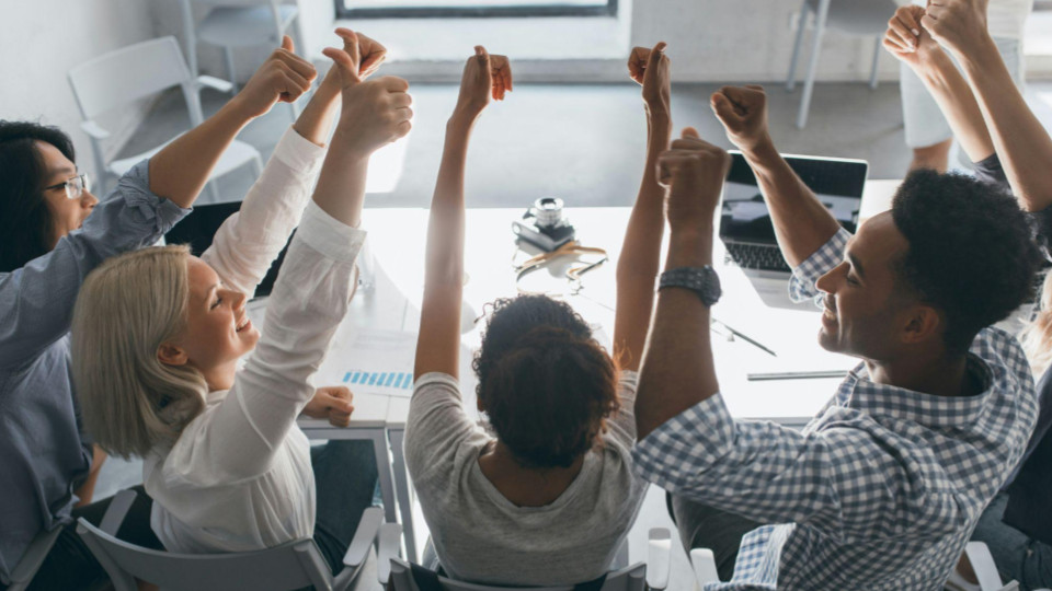 Grupo de pessoas em uma reunião de trabalho sentadas ao redor de uma mesa, levantando as mãos em um momento de celebração ou aprovação de uma decisão.
