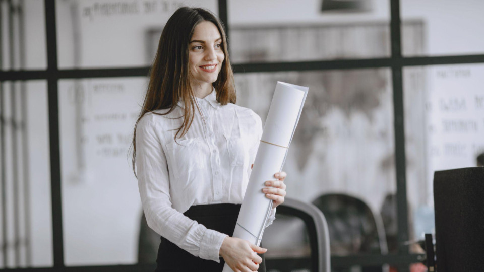 Uma mulher sorridente, vestindo camisa branca e saia preta, está em um ambiente corporativo moderno enquanto segura um projeto ou documento enrolado. Ao fundo, há divisórias de vidro, um quadro com informações e elementos de escritório.