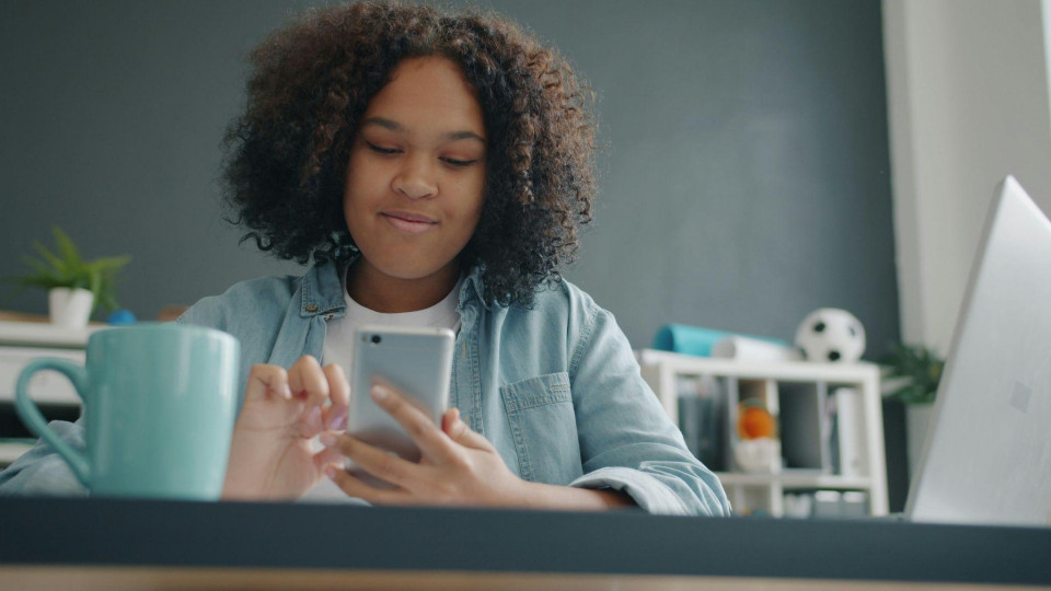 Colaboradora utilizando um smartphone enquanto trabalha em uma mesa de escritório, representando o uso de soluções digitais para gestão de benefícios, comunicação e praticidade no ambiente corporativo.