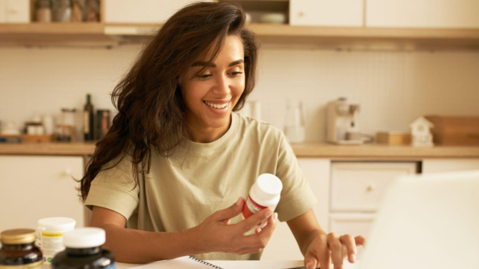 A imagem mostra uma mulher sorridente em uma cozinha clara e bem iluminada em tons quentes e amadeirados. Detalhes principais: Pessoa: Uma mulher jovem com cabelo escuro e ondulado, vestindo uma camiseta casual na cor verde-oliva clara. Ela está olhando para baixo com um sorriso expressivo segurando medicamentos.