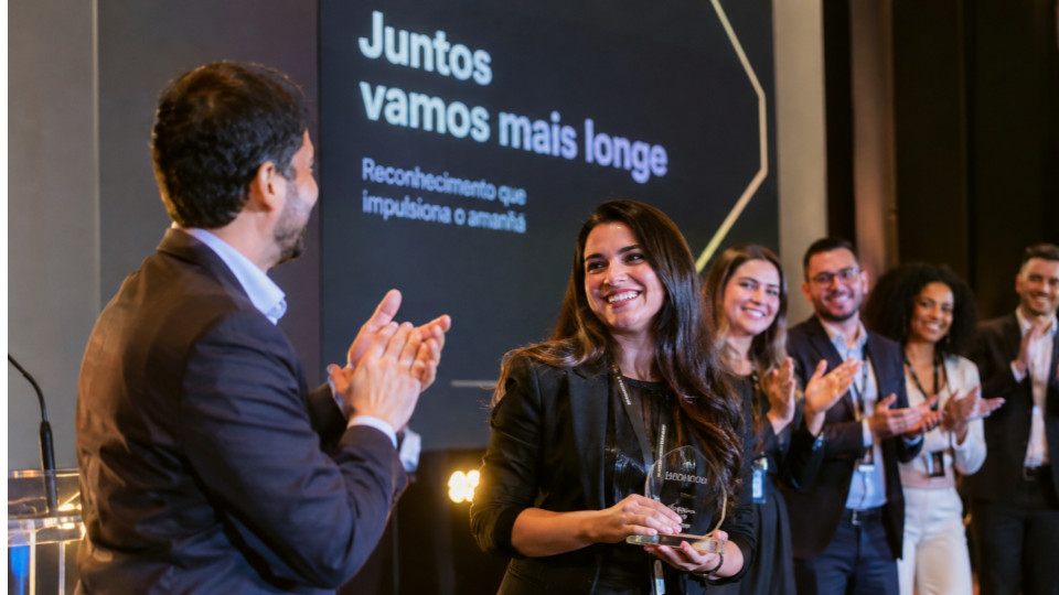 Fotografia realista de uma cerimônia de premiação corporativa em uma empresa moderna, realizada em um ambiente elegante, com iluminação profissional e palco com painel de fundo sofisticado. No centro da cena, uma funcionária sorridente recebe um troféu de reconhecimento, enquanto um colega aplaude e outros profissionais, vestidos com trajes sociais e executivos, comemoram a conquista ao fundo.
