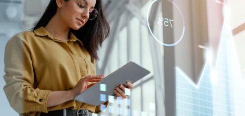 A imagem mostra uma mulher jovem analisando dados em um tablet, em um ambiente moderno e iluminado, com elementos gráficos digitais flutuando ao seu redor.