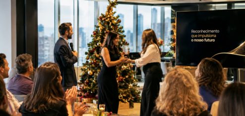 Fotografia realista de uma premiação de fim de ano em uma empresa moderna, realizada em um ambiente corporativo elegante, com amplas janelas de vidro e vista para a cidade. No centro da cena, uma colaboradora recebe um troféu de reconhecimento de outra profissional, enquanto um apresentador segura um microfone e os colegas assistem e aplaudem.