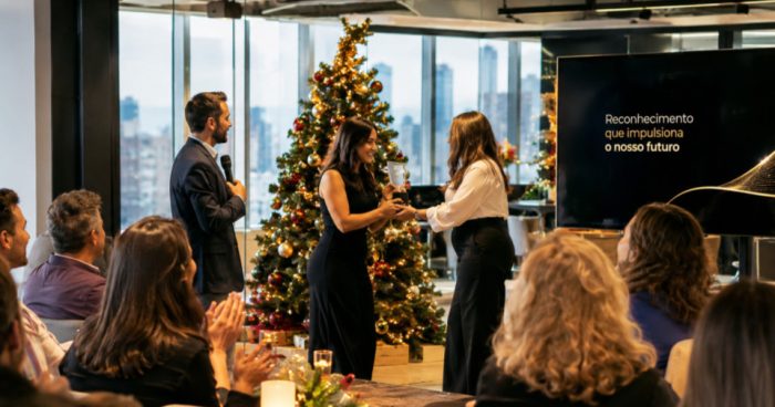 Fotografia realista de uma premiação de fim de ano em uma empresa moderna, realizada em um ambiente corporativo elegante, com amplas janelas de vidro e vista para a cidade. No centro da cena, uma colaboradora recebe um troféu de reconhecimento de outra profissional, enquanto um apresentador segura um microfone e os colegas assistem e aplaudem.