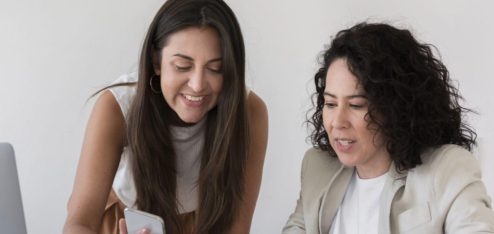 A imagem mostra duas mulheres em um ambiente de trabalho, sentadas ou inclinadas sobre uma mesa, analisando algo em um smartphone.