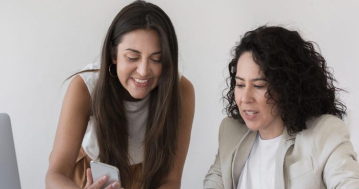 A imagem mostra duas mulheres em um ambiente de trabalho, sentadas ou inclinadas sobre uma mesa, analisando algo em um smartphone.