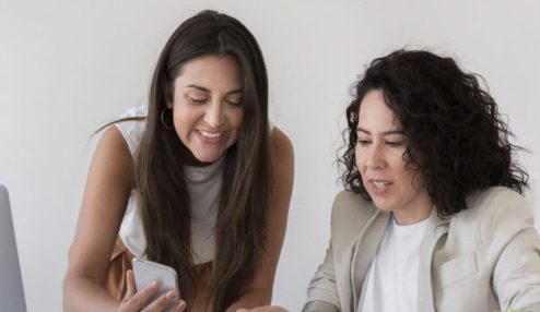 A imagem mostra duas mulheres em um ambiente de trabalho, sentadas ou inclinadas sobre uma mesa, analisando algo em um smartphone.