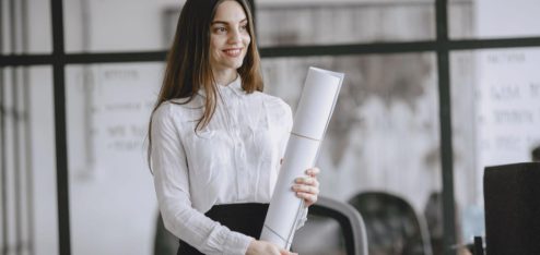 Uma mulher sorridente, vestindo camisa branca e saia preta, está em um ambiente corporativo moderno enquanto segura um projeto ou documento enrolado. Ao fundo, há divisórias de vidro, um quadro com informações e elementos de escritório.