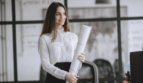 Uma mulher sorridente, vestindo camisa branca e saia preta, está em um ambiente corporativo moderno enquanto segura um projeto ou documento enrolado. Ao fundo, há divisórias de vidro, um quadro com informações e elementos de escritório.