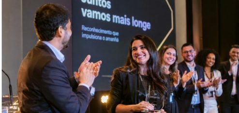 Fotografia realista de uma cerimônia de premiação corporativa em uma empresa moderna, realizada em um ambiente elegante, com iluminação profissional e palco com painel de fundo sofisticado. No centro da cena, uma funcionária sorridente recebe um troféu de reconhecimento, enquanto um colega aplaude e outros profissionais, vestidos com trajes sociais e executivos, comemoram a conquista ao fundo.