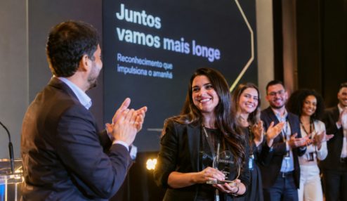 Fotografia realista de uma cerimônia de premiação corporativa em uma empresa moderna, realizada em um ambiente elegante, com iluminação profissional e palco com painel de fundo sofisticado. No centro da cena, uma funcionária sorridente recebe um troféu de reconhecimento, enquanto um colega aplaude e outros profissionais, vestidos com trajes sociais e executivos, comemoram a conquista ao fundo.