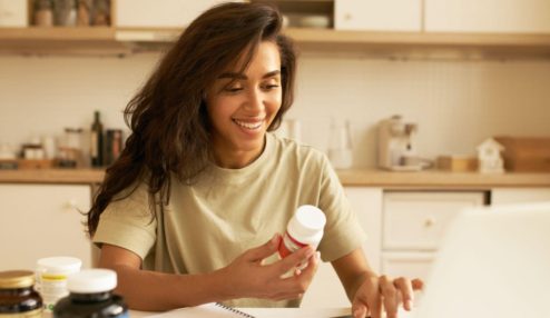 A imagem mostra uma mulher sorridente em uma cozinha clara e bem iluminada em tons quentes e amadeirados. Detalhes principais: Pessoa: Uma mulher jovem com cabelo escuro e ondulado, vestindo uma camiseta casual na cor verde-oliva clara. Ela está olhando para baixo com um sorriso expressivo segurando medicamentos.
