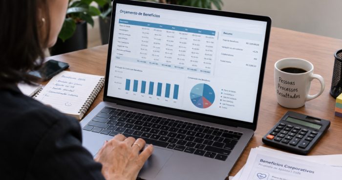 Profissional de RH analisando uma planilha de orçamento de benefícios em um notebook, com calculadora, café e documentos sobre a mesa.