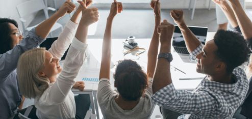 Grupo de pessoas em uma reunião de trabalho sentadas ao redor de uma mesa, levantando as mãos em um momento de celebração ou aprovação de uma decisão.