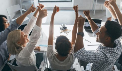 Grupo de pessoas em uma reunião de trabalho sentadas ao redor de uma mesa, levantando as mãos em um momento de celebração ou aprovação de uma decisão.
