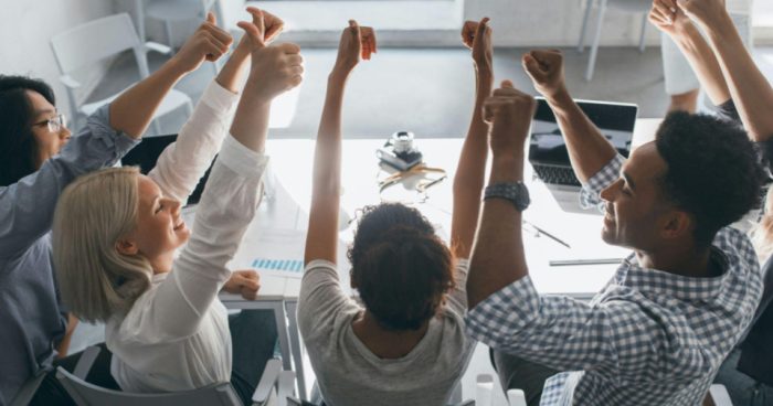Grupo de pessoas em uma reunião de trabalho sentadas ao redor de uma mesa, levantando as mãos em um momento de celebração ou aprovação de uma decisão.