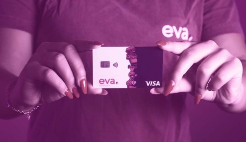 Pessoa com camiseta da 'Eva'. Nas mãos da pessoa, de unhas vermelhas, e em frente ao peito, está um cartão vale-alimentação e vale-refeição Eva.
