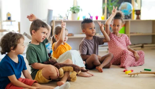 Crianças felizes levantando as mãos em sala de aula, participando de atividade lúdica com brinquedos e objetos coloridos. Ambiente acolhedor e educativo.