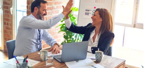A imagem mostra dois profissionais em um ambiente de escritório celebrando um momento de sucesso com um entusiasmado "high five" (toca aqui).