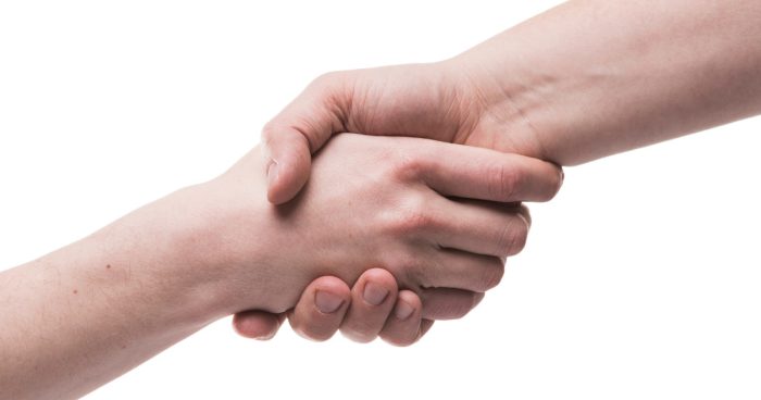 A imagem mostra duas pessoas se cumprimentando com um aperto de mão. A mão esquerda, em primeiro plano, está sendo segurada pela mão direita, que está em segundo plano.